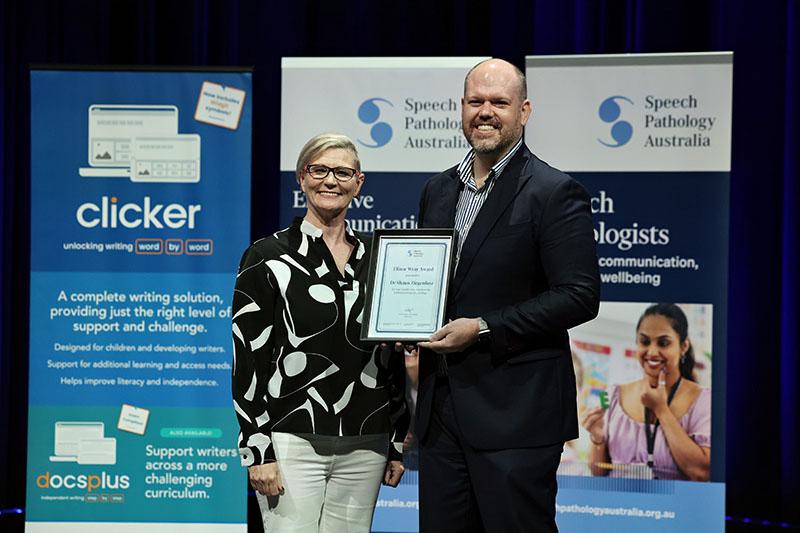 This is a photo of Dr Shaun Ziegenfusz holding a Elinor Wray Award plaque as he poses with Emma Power to his left at the SPA 2026 Conference. Emma has short blonde hair and is wearing a black and white shirt with white pants. Shaun is wearing a black suit with a striped shirt.