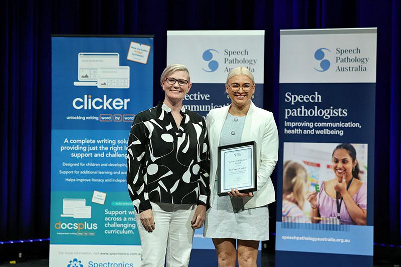 This is a photo of Dr Clancy Conlon holding a Service and Impact Award plaque as she poses with Emma Power to her left at the SPA 2026 Conference. Emma has short blonde hair and is wearing a black and white shirt with white pants. Clancy has blonde hair tied back and is wearing a white blazer over a grey dress.