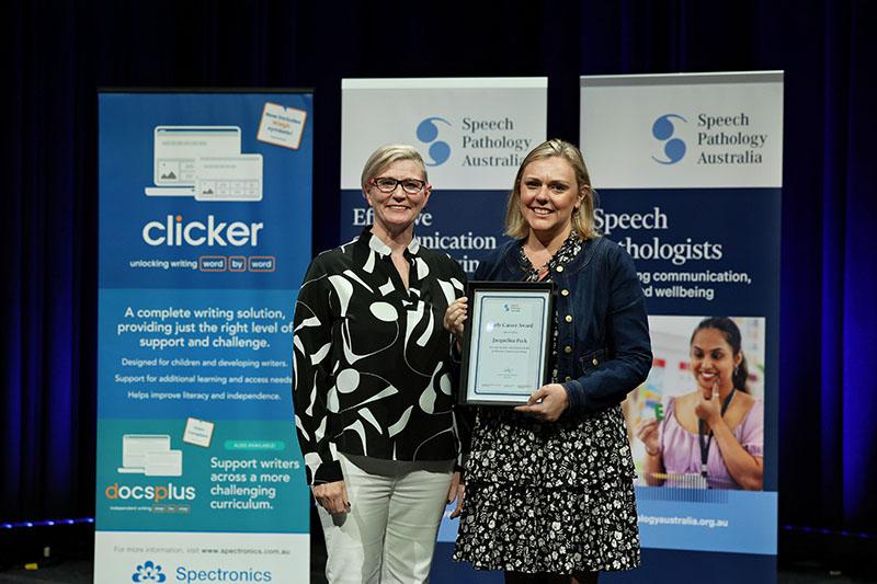 This is a photo of Jacqueline Peck holding an Early Career Award plaque as she poses with Emma Power to her left at the SPA 2026 Conference. Emma has short blonde hair and is wearing a black and white shirt with white pants. Jacqueline has mid-length blonde hair and is wearing a denim jacket over a black floral dress.