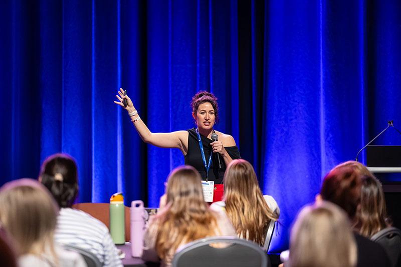 This is a photo of a woman presenting in front of a crowd. Rebecca is pointing upwards as she speaks.