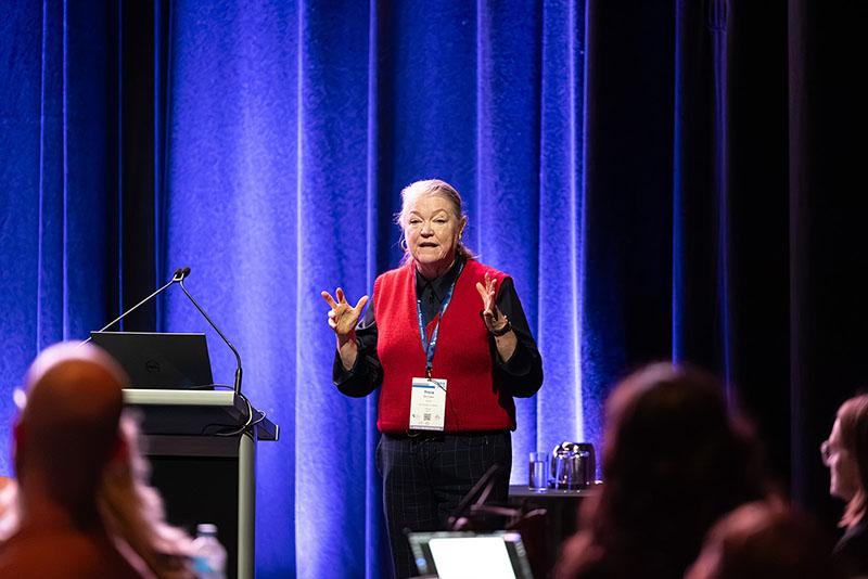 This is a photo of a woman presenting in front of a crowd. Tricia is holding up her hands as she speaks in detail.