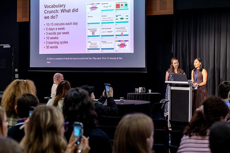 This is a photo of 2 women presenting behind a podium to an audience. There is a projector in the back that says, ‘Vocabulary Crunch: What did we do?’