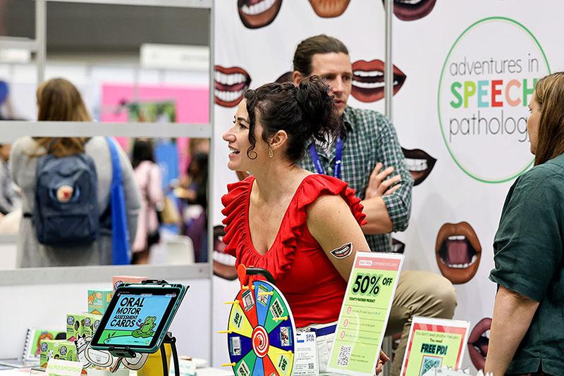 This is a photo of 2 women and 1 man at the Adventures in Speech Pathology Booth. The booth has a number of items on the table such as an ipad with a game on it, a wheel of fortune and cards.