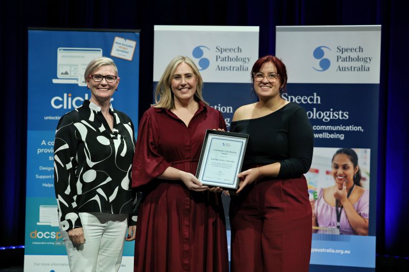 This is a photo of 2 women wearing maroon and black coloured clothing holding a Community Contribution Award plaque together. Emma Power stands to their left. They are on stage at the SPA 2026 Conference. Emma has short blonde hair and is wearing a black and white shirt with white pants.