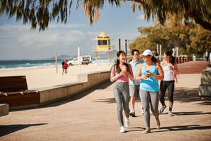 Two people wearing athletic wear, walking along a beachside path, each holding a coffee, others walking behind them. Source: Destination Gold Coast