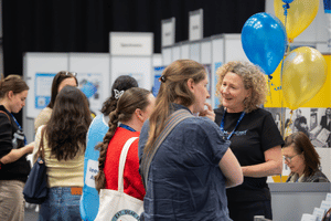 Attendees speaking to exhibitors, with gold and blue balloons at one of the stands.