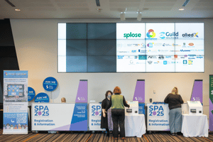 SPA 2025 Conference branded registration and information desk, with staff and attendees chatting in front of it. A large screen behind displays a list of sponsors and exhibitors.