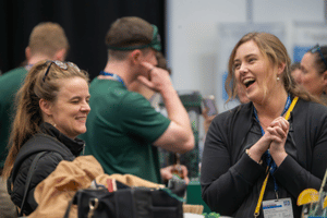 Two attendees in conversation, one laughing with hands clasped. Other Attendees can be seen in the background. 
