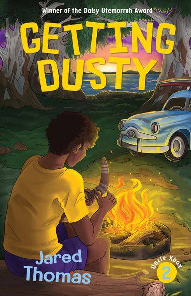 This is the cover for ‘Getting Dusty’. Written by Jared Thomas. The cover shows a boy holding a boomerang as he sits over a fire in the outback with a car in the background.