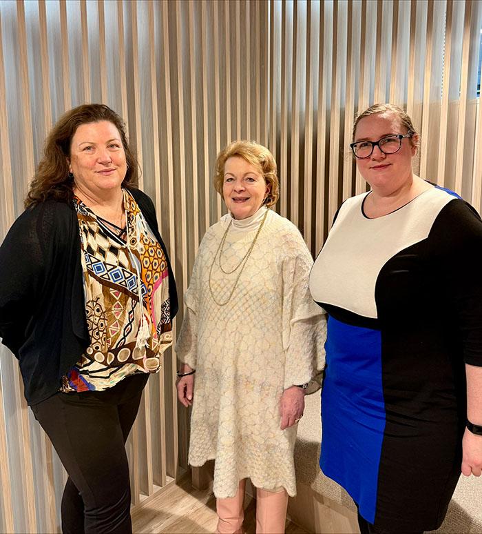Three people standing indoors in front of a wall of vertical timber slats. One person wears a patterned top and dark cardigan, another wears a light-coloured textured sweater with a long necklace, and the third wears a black, white and blue dress with glasses. The group is posing for a photo in a modern office or meeting space. Three people standing indoors in front of a wall of vertical timber slats. One person wears a patterned top and dark cardigan, another wears a light-coloured textured sweater with a long necklace, and the third wears a black, white and blue dress with glasses. The group is posing for a photo in a modern office or meeting space.