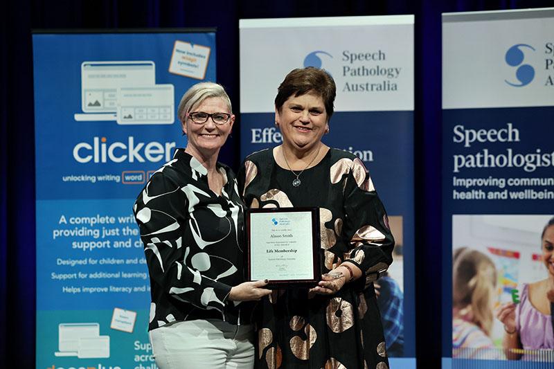 This is a photo of Alison Smith holding a Life Membership plaque as she poses with Emma Power to her left at the SPA 2026 Conference. Emma has short blonde hair and is wearing a black and white shirt with white pants. Alison has short brown hair and is wearing a black and bronze dress. 
